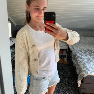 Beige stickad kofta från XOXO Friends - Beige stickad väst från XOXO Friends med grovstickad struktur och knappar framtill. Modellen är croppad och har breda muddar vid ärmslut och nederkant. Perfekt att slänga över en t-shirt för en chill look. Har nån lite fläck (se bild 4) men den kanske går bort i tvätten.