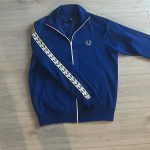 Blå track jacket från Fred Perry - Säljer en snygg blå track jacket från Fred Perry med vita detaljer och dragkedja. Jackan har långa ärmar med ett mönstrat band längs ärmarna och en broderad logga på bröstet. Perfekt för en skön look!