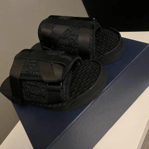 Svarta dior alpha slides - Snygga svarta sandaler från Dior med breda remmar och diskret Dior-monogram över hela ovandelen. Sandalerna har platt sula och mönstrad fotbädd för extra komfort. Perfekta för en clean och modern look.