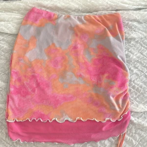 Färgglad kortkjol från Wild Fable XS - Superfin kortkjol från Wild Fable i mesh med vågiga kanter och dragsko på sidan. Kjolen har ett färgstarkt mönster i rosa, orange och grått, samt dubbla lager för en cool effekt. Perfekt för dig som vill sticka ut i sommar!