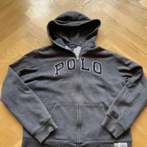 Grå hoodie från Polo Ralph Lauren  - Mörkgrå hoodie från Polo Ralph Lauren i storlek XL(barnstorlek)  S (herrstorlek)med stor broderad POLO-text i blått och vitt på bröstet. Tröjan har huva, dragkedja hela vägen och känguruficka. Mjuk insida och klassisk collegekänsla. Perfekt för chill dagar.