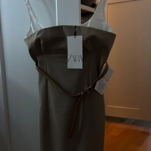 Beige klänning med bälte från Zara - Super fin och elegant beige klänning från Zara med smala axelband i vitt och ett brunt bälte i midjan. Helt ny med prislappen kvar, säljer tyvärr för den ej har kommit till användning. 