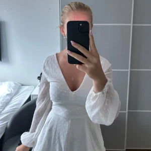 Vit kortklänning med volanger och knyt - Superfin vit kortklänning  med v-ringning, långa transparenta ärmar och volanger i kjolen. Prickigt mönster i tyget och knytband i midjan ger en romantisk vibe. Perfekt för sommarens alla tillfällen!