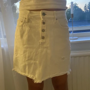 Vit jeanskjol med knappar och rå kant - Säljer en vit jeanskjol med snygga slitningar, rå kant nertill och silverfärgade knappar framtill. Kjolen har fem fickor och en rak passform som sitter skönt på höften. Perfekt för dig som gillar en avslappnad och trendig stil.
