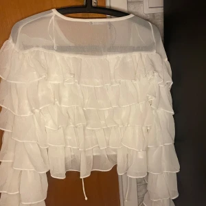 Vit volangig bolerotopp från Gina Tricot - Superfin vit bolerotopp från Gina Tricot med massor av volanger och transparenta detaljer. Toppens material är lätt och luftig polyester, och den har långa ärmar med volang hela vägen. Perfekt för dig som vill sticka ut med en romantisk och trendig look.