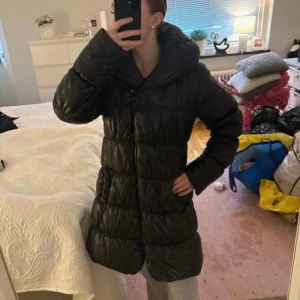 Svart lång pufferjacka med huva - Säljer en svart lång pufferjacka med stor huva och quiltad design. Jackan är riktigt fluffig och har dragkedja framtill. Perfekt för kalla dagar när du vill hålla dig varm och ändå se snygg ut. Ganska glansig. Originalpris 1200kr