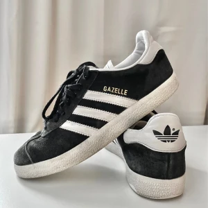 Adidas Gazelle Svart - Klassiska Adidas Gazelle sneakers i svart mocka med vita detaljer och de ikoniska tre ränderna på sidorna. Låg modell med snörning och vit sula. Adidas logga på hälen och guldtext på sidan. Perfekta för en clean och sportig stil.