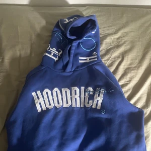 Blå HOODRICH hoodie med brodyr - Säljer en blå hoodie från HOODRICH med stor vit brodyr på bröstet och coola broderade detaljer på huvan. På ryggen finns vit text med motiverande citat. Hoodien har känguruficka och dragsko i huvan. Perfekt för dig som gillar streetwear vibes.