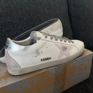 Golden Goose vita sneakers med silverdetaljer - Snygga vita sneakers från Golden Goose med grå snörning, silverfärgad häl och ikonisk stjärna i mocka på sidan. Skorna har en platt sula och detaljer i både skinn och mocka för en cool, urban look.