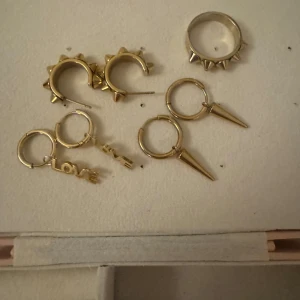 Edblad Guldiga örhängen & ring med spikes - Säljer ett set med tre par guldiga örhängen och en matchande ring. Örhängena har coola detaljer: ett par med spetsiga hängen, ett par med texten LOVE och ett par med små spikes. Ringen är bred och dekorerad med spikes för en extra vibe. Perfekt för dig som gillar Sthlm stil! ( Ringen storlek 17,5/ obs peak örhängena har ingen plupp ibak )