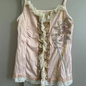 Rosa linnetopp med spets från Saint Tropez - Supersöt ljusrosa linnetopp från Saint Tropez med tunna axelband, volangdetaljer och spetskant nertill. Framsidan har en stor broderad blomma, dekorativa band och rynkade partier i beige och vitt. Perfekt för dig som gillar romantiska och unika detaljer. Storlek M men lite mindre