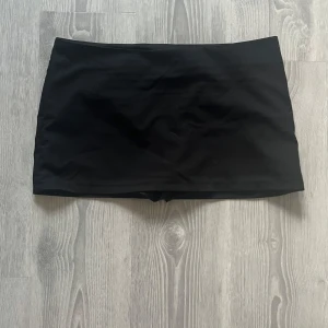 Svart kortkjol med inbyggda shorts - Säljer en svart, tight och enkel minikjol med inbyggda shorts under. Kjolen har en clean och stilren look, perfekt för dig som vill ha något basic men ändå praktiskt. Kjolen stängs med en dragkedja på sidan och är inte så stretchig.