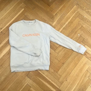 Ljusgrå Calvin Klein Jeans sweatshirt - Snygg ljusgrå sweatshirt från Calvin Klein Jeans med orange logga framtill och detalj på ärmen. Tröjan har rund hals, långa ärmar och ribbade muddar. Perfekt för en chill och stilren look.