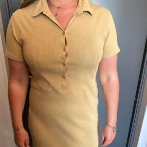 Gul ribbad kortklänning med krage - Snygg gul ribbad klänning med korta ärmar och klassisk krage. Klänningen har knappar framtill och en figurnära passform som ger en clean och stilren look.