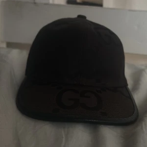 Svart keps från Gucci i skinn - Snygg svart keps från Gucci med GG-logga framtill. Kepsen har böjd skärm i präglat skinn och justerbart spänne bak i läder. Klassisk och stilren design som lyfter vilken outfit som helst.