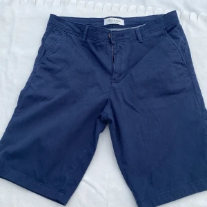 Marinblå shorts från Stock & Hank - Klassiska marinblå shorts från Stock & Hank med normal passform. De har fickor fram och bak, bälteshällor och stängs med dragkedja och knapp. Perfekta för sommardagar och enkla att matcha med t-shirt eller skjorta.