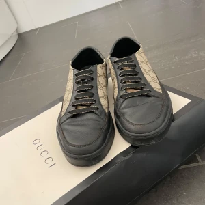 Gucci sneakers med GG-mönster - Snygga sneakers från Gucci med klassiskt GG-monogram i beige och svarta detaljer i skinn. Skorna har svarta snören och en platt sula med Gucci-logga undertill. Perfekta för dig som vill ha en lyxig och tidlös look med streetkänsla.