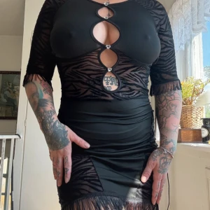 Svart kort zebra-klänning med cutouts - Snygg svart kort klänning med zebramönster och genomskinliga meshpartier. Klänningen har flera cutouts framtill med glittriga detaljer, fransar vid ärmslut och nederkant samt trekvartsärm. Tight passform och cool design som sticker ut.