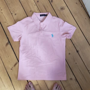 Rosa pikétröja från Polo Ralph Lauren - Snygg ljusrosa pikétröja från Polo Ralph Lauren med klassisk krage och korta ärmar. Tröjan har blå broderad logga på bröstet och är gjord i mjuk bomull. Perfekt för en clean och fräsch look.