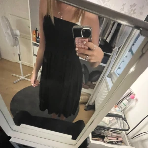 Svart axelbandslös klänning - Snygg svart klänning med axelbandslös design och lätt utställd kjol. Perfekt för dig som gillar en stilren look med lite edge. Klänningen har en klassisk passform och är enkel att styla med smycken eller en cool jacka.