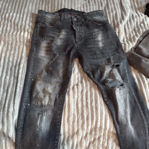 Svarta distressed jeans från Dsquared2 - Säljer ett par svarta jeans från Dsquared2 med slitna detaljer, hål och färgstänk för en riktigt edgy look. Jeansen har fem fickor, normal passform och är tillverkade i klassisk denim. Perfekta för dig som gillar streetstyle och vill sticka ut.