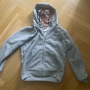 Grå hoodie från Burberry med dragkedja - Snygg grå hoodie från Burberry i 100% bomull. Klassisk modell med huva, snörning och dragkedja framtill. Fodret i huvan har Burberrys ikoniska rutmönster i beige, svart och rött. Två stora fickor framtill och ribbade muddar vid ärmslut och midja. Sparsamt användt utan några slitage. 