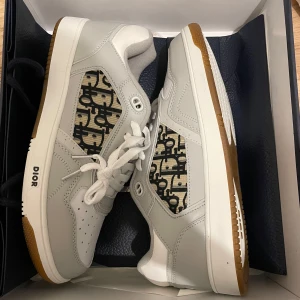 Dior B23 sneakers med monogram - Snygga Dior B23 sneakers i vitt och grått skinn med klassiskt Dior-monogram i svart på beige textilpanel. Skorna har vit snörning, perforerad tå och gummisula i vitt och brunt. Perfekt för dig som vill sticka ut med exklusiva detaljer och modern design.