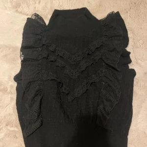 Svart stickad volangtröja med spets - Supersnygg svart stickad tröja med höghalsad krage och dekorativa volanger i spets längs axlarna och framtill. Tröjan har en feminin och trendig look med coola detaljer som ger extra edge till outfiten. Mycket bra skick. Passar xs-L