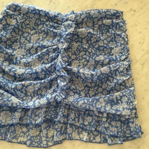 Ny Blåvit blommig volangkjol - Supersöt kort kjol i blått och vitt med blommigt mönster och volanger. Kjolen har rynkade detaljer framtill och är gjord i ett lätt, luftigt material som passar perfekt till varma dagar. Perfekt för dig som gillar romantisk och somrig stil!