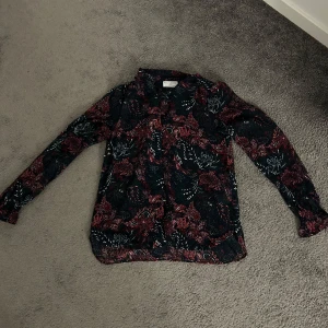 Blommig blus från Holly & White - Mörkblå blus med röda och vita blommor från Holly & White. Långärmad modell med lätt transparent tyg och diskreta knappar framtill. Blusen har avslappnad passform och elastiska ärmslut för en snygg siluett.