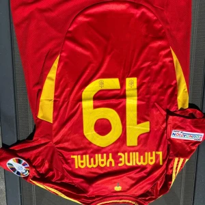 Spansk landslagströja Yamal 19 Adidas - Röd fotbollströja från Addidas med gula detaljer och nummer 19, Lamine Yamal på ryggen. Officiella märken för Spaniens landslag och turneringspatchar på ärmarna. Klassisk rund hals och korta ärmar. Tillverkad i lätt och ventilerande polyester.
