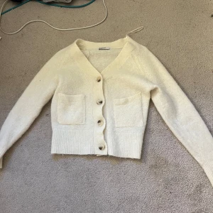 Krämvit kofta i alpackablandning XS - Supermjuk och fluffig krämvit kofta från Cubus i alpackablandning. Koftan har v-ringning, stora knappar framtill, två fickor och ribbade muddar. Perfekt för lager-på-lager och ger en chill men ändå stilren vibe.