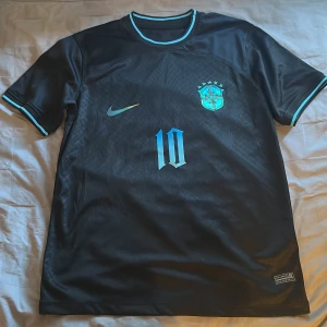 Svart Brasilien fotbollströja Nike - Snygg svart Brasilien fotbollströja från Nike med turkosa detaljer och nummer 10 på både fram- och baksida. CBF-emblem på bröstet, turkosa ränder vid krage och ärmslut. Tillverkad i lätt och ventilerande material, perfekt för match eller träning.