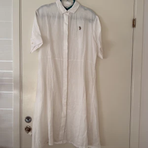 Vit linneskjortklänning U.S. Polo Assn. - Vit skjortklänning från U.S. Polo Assn. i luftig linne med klassisk krage, korta ärmar och knäppning framtill. Broderad logga på bröstet ger en clean och sportig vibe. Perfekt för varma dagar när du vill vara både chill och snygg.