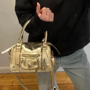 Guldig city väska - Cool guldig handväska. Väskan har både handtag och justerbar axelrem, dragkedja upptill och en liten ficka med dragkedja framtill. 