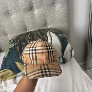Burberry  keps  - Snygg keps från Burberry med klassiskt rutigt mönster i beige, svart, vitt och röda linjer. Justerbar baksida med svart spänne och Burberry London England-logga. Perfekt accessoar för att lyfta din outfit med en lyxig touch. En knapp är av men det syns inte riktigt då jag använt svart eltejp som gör det svårt att synas 