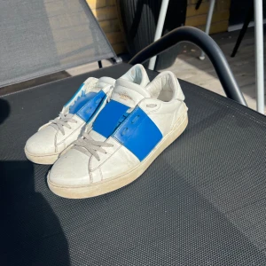 Valentino Open sneakers vit/blå - Snygga Valentino Open sneakers i vitt skinn med en bred blå läderdetalj över mitten. Klassisk rund tå, vita snören och diskret logga på plösen. Perfekta för dig som gillar statement-detaljer och exklusiv känsla.