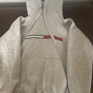 Grå hoodie Tommy Hilfiger  - Snygg ljusgrå hoodie från Tommy Hilfiger med klassisk logga och ränder i vitt, blått och rött på bröstet. Tröjan har huva med snörning, magficka och långa ärmar. Perfekt för en chill och avslappnad stil.