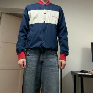 Adidas blå och röd bomberjacka - Retro bomberjacka från Adidas i marinblått med bred vit panel och röda detaljer. Stor röd 72 på ryggen och Adidas-logga fram och bak. Jackan har knappar, ribbade muddar och krage, samt två fickor framtill. Perfekt för dig som gillar sportig vintage-stil. Lätt missfärgning på den vita delen men det går att se över.