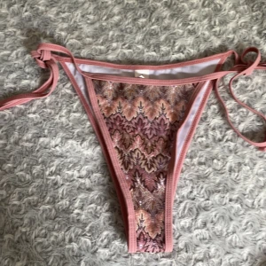 Rosa bikiniunderdel med spetsmönster M - Supersnygg bikiniunderdel i rosa med spetsliknande mönster. Knytband i sidorna för perfekt passform. Materialet är 100% polyester. Aldrig använda. 