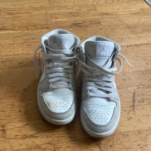 Nike Air Jordan 1 Mid i vitt och grått - Nike Air Jordan 1 Mid sneakers i vitt och ljusgrått skinn och mocka, med klassisk rosa Swoosh på sidan. Hög modell med snörning och perforerad tå för extra ventilation. Ikonisk Air Jordan-logga på sidan och mjuk vaddering runt ankeln.