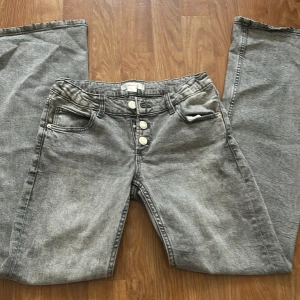 Grå jeans med knappar från Gina Tricot - Snygga grå jeans från Gina Tricot med raka ben och flera silverfärgade knappar framtill. Jeansen har klassiska fem fickor, bälteshällor och en cool tvättad look. Perfekta för dig som gillar en avslappnad men trendig stil.