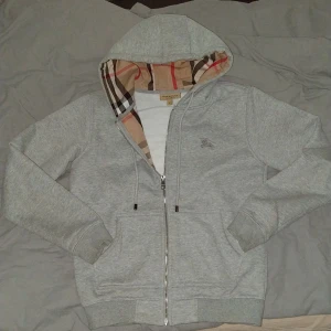 Burberry hoodie grå - Snygg grå hoodie från Burberry med klassiskt rutigt foder i huvan och broderad logga på bröstet. Tröjan har dragkedja framtill, snörning i huvan och två fickor. Storlek S