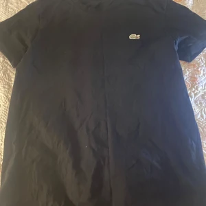 Svart t-shirt från Lacoste - En klassisk svart t-shirt från Lacoste med den ikoniska krokodilloggan broderad på bröstet. T-shirten har rund halsringning och korta ärmar. Tillverkad i mjuk bomull som känns skön mot huden. Perfekt för en clean och stilren look.