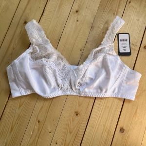 Vit spetsblus från Trofé 105B - Säljer en vit blus från Trofé med vacker spets upptill och breda axelband. Blusen har en klassisk form och är tillverkad i ett mjukt, glansigt material med fina spetsdetaljer som ger en romantisk vibe. Perfekt för dig som gillar stilrena och feminina plagg.