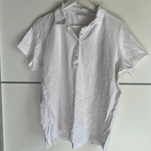 Vit piké t-shirt från Isolde XL - Klassisk vit piké t-shirt från Isolde i storlek XL. T-shirten har korta ärmar, krage och knäppning med fyra knappar framtill. Tillverkad i mjuk bomull, perfekt för en clean och enkel stil.