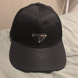 Svart keps från Prada i nylon - Snygg svart keps från Prada med klassisk böjd skärm och ikonisk metallogo framtill. Justerbar rem baktill i svart läder med präglad Prada Milano-text. Kepsen är tillverkad i glansig nylon som ger en lyxig känsla och modern look.Tar även emot byten helst Gucci keps original 