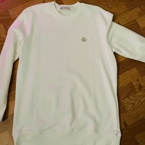 Vit sweatshirt från Moncler, storlek L - Säljer en stilren vit sweatshirt från Moncler i storlek L. Tröjan har rund halsringning, lång ärm och en diskret Moncler-logga på bröstet. Tillverkad i mjuk bomull för en clean och fräsch look. Perfekt för dig som gillar minimalistisk stil.