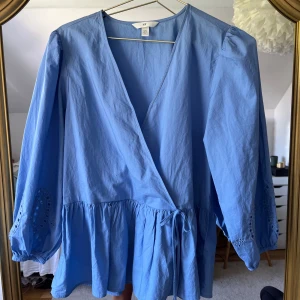 Blå omlottblus med broderi från H&M - Supersöt ljusblå omlottblus från H&M med knyt i midjan och volang nedtill. Blusen har långa ärmar med broderade detaljer och elastiska muddar. Sparsamt använd💕