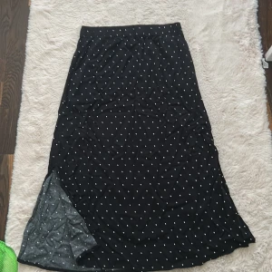 Svart prickig långkjol från 157 - Svart långkjol från LSL med vita prickar över hela tyget. Kjolen har resår i midjan för en bekväm passform och är tillverkad i ett mjukt, lätt material. Perfekt för dig som gillar en klassisk men ändå lekfull stil.